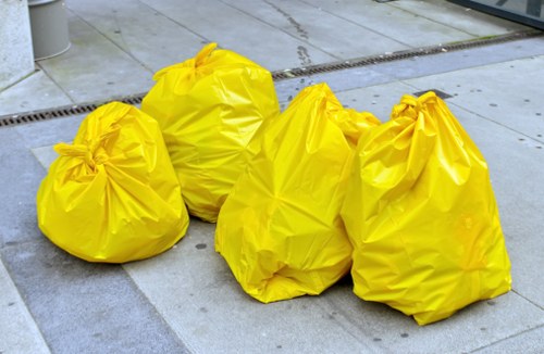 Four large yellow rubbish bags made of thick, glossy plastic are stacked on a paved outdoor surface. The bags are loosely gathered at the top, with some appearing slightly overfilled, and they have a smooth, shiny finish that reflects ambient light. One bag on the left is positioned slightly in front of the others, which are aligned behind it. The setting appears to be an urban or commercial area, with a concrete or stone pavement and a metal drainage grate running across the ground behind the bags. In the background, part of a building or wall is visible, suggesting the bags are placed outside a business or residential property. The presence of these rubbish bags aligns with the services offered by Commercial Waste Removal Edmonton, which specialises in the collection and disposal of waste materials in the local area, including Edmonton and nearby regions.