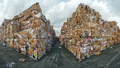 Two large stacks of compressed cardboard and paper waste are situated outdoors on a paved surface, with each pile towering several meters high and extending into the distance. The waste material appears to be a mix of brown kraft paper, corrugated cardboard, and flattened paperboards, with some coloured paper and printed materials visible sporadically throughout. The stacks are arranged in parallel, creating a narrow pathway between them, and their surfaces show a combination of rough, crinkled textures from the compressed paper, interspersed with smoother, folded edges. The background features a cloudy sky, casting diffuse lighting across the scene, while the surrounding area appears to be an industrial or waste management site, possibly part of waste collection or recycling operations linked to a local rubbish removal service such as Commercial Waste Removal Edmonton. The overall environment emphasizes a formal storage and handling of bulky paper waste material, characteristic of commercial waste management activities in the Edmonton area or similar localities.