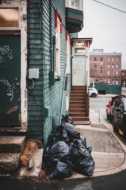 An outdoor scene featuring a pile of mixed rubbish and waste materials accumulated on a gravel surface beside a low stone wall. The waste includes black plastic bags, a yellow plastic container, a ben