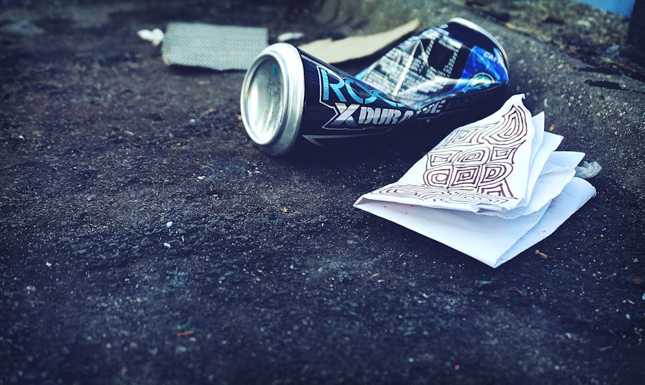 A discarded aluminium can lies on a dark, textured pavement, partially crushed with a shiny silver top and black and blue branding visible. Next to the can, there is a crumpled piece of white paper wi