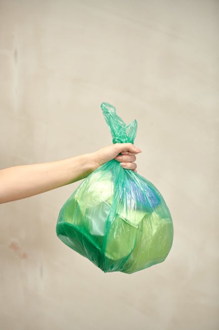 Two waste collection workers wearing dark green uniforms with reflective orange stripes are engaged in rubbish removal on a city street, loading a large blue bag of waste into a waste management vehic